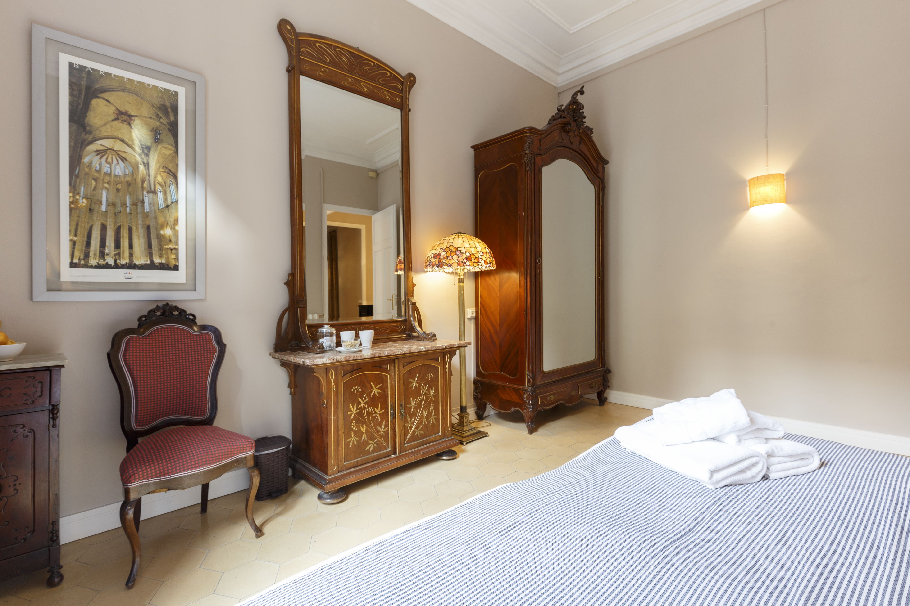 A hotel room with a bed, a chair, a dresser, a mirror, and a wall-mounted lamp.