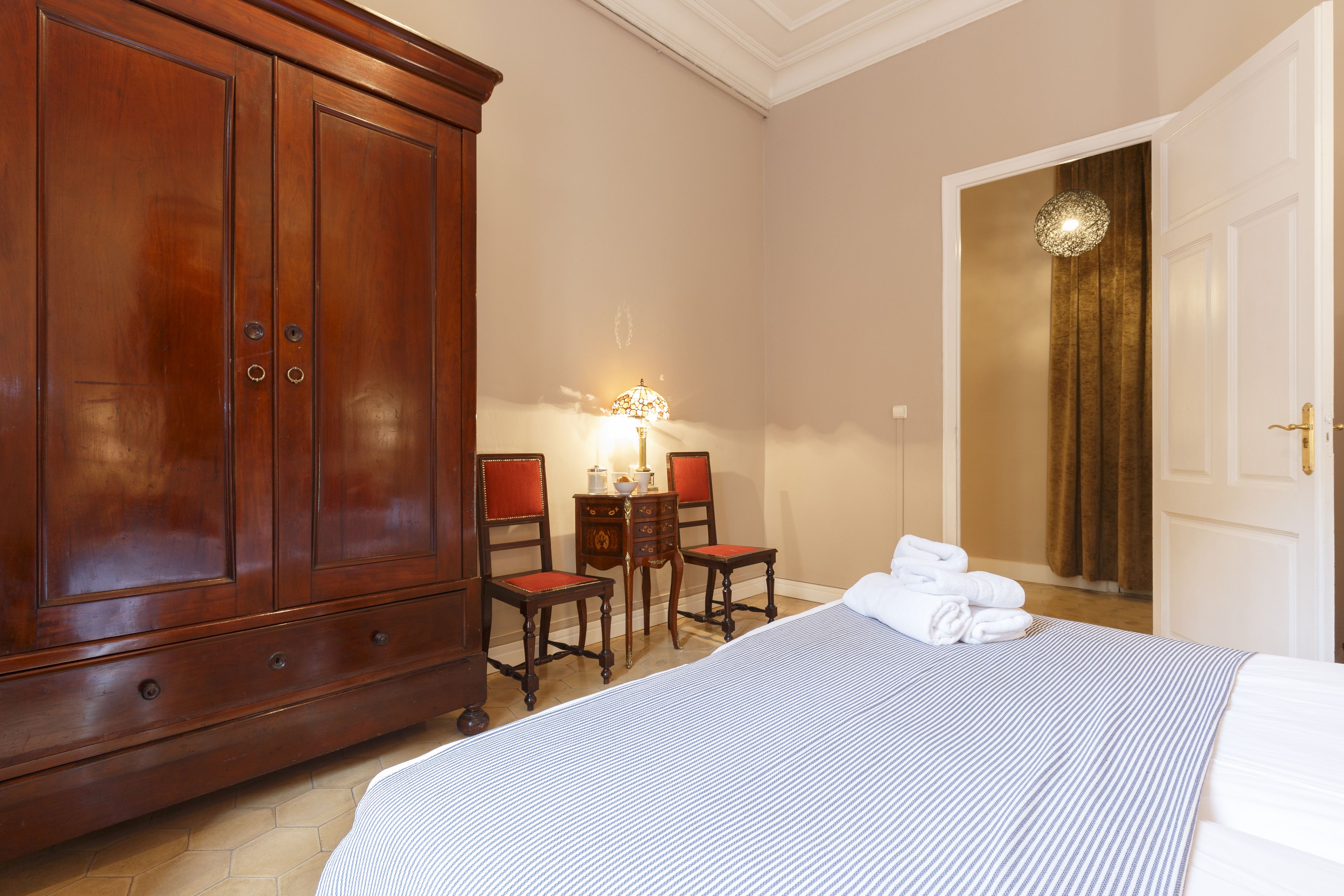 A hotel room with a large wooden wardrobe, a bed with a striped bedspread, a desk with two chairs, and a lamp.