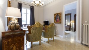 Une pièce avec un lustre, une table surmontée d’une lampe, deux fauteuils richement décorés, un tableau encadré et un miroir.