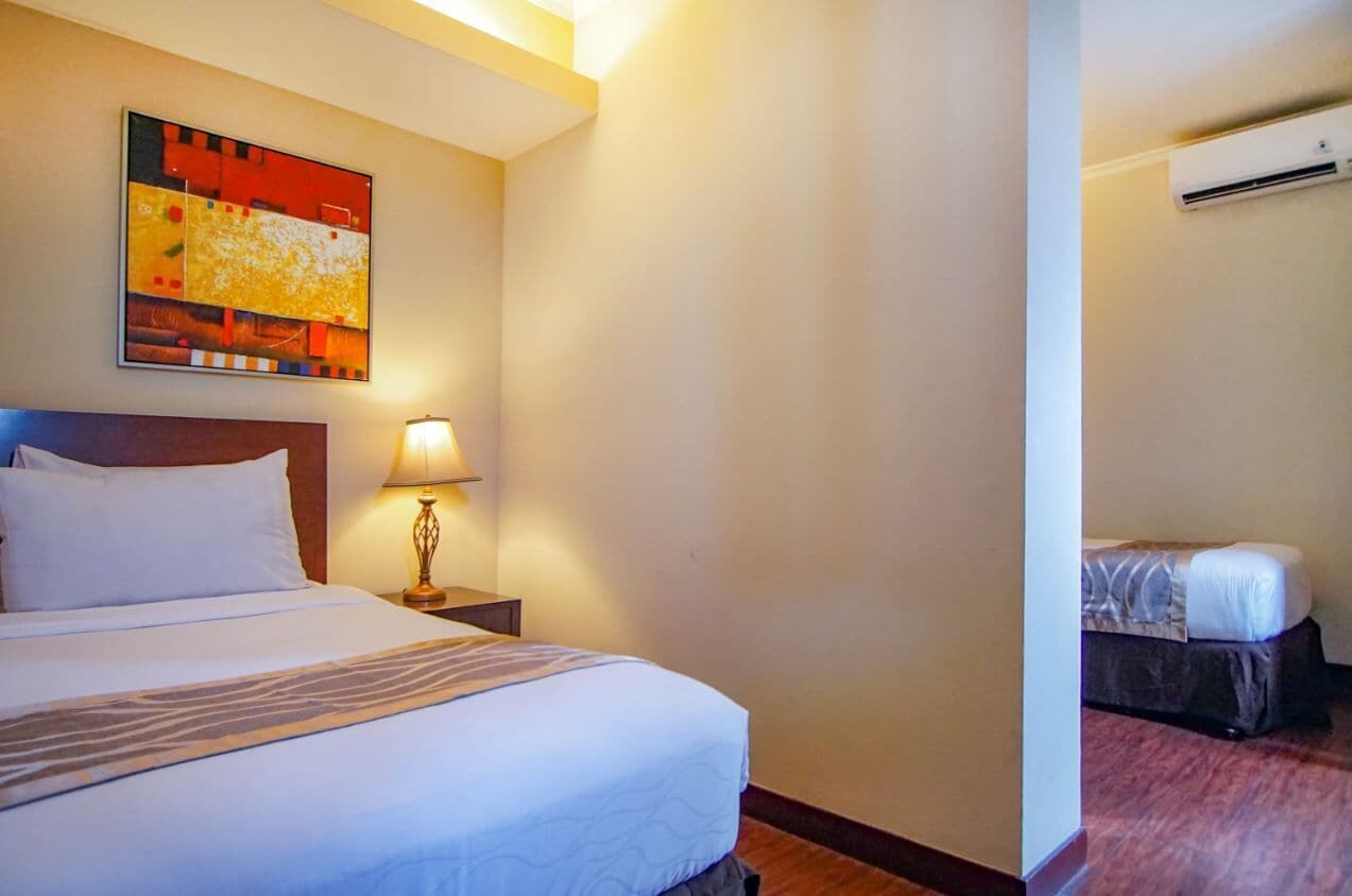 A hotel room with a large bed, a bedside lamp, a small table, a wall-mounted air conditioner, and a painting above the bed.