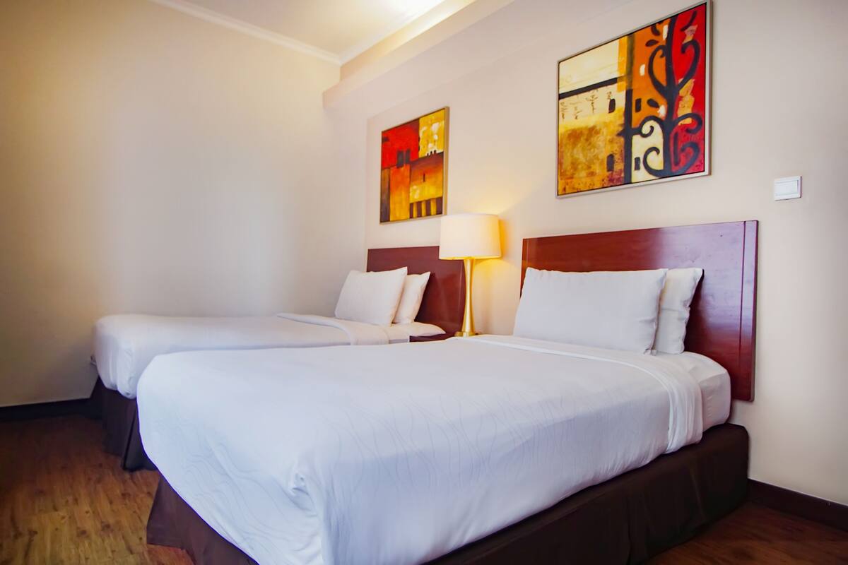 Une chambre d’hôtel avec deux lits simples, une tête de lit en bois, une table de chevet avec une lampe et des œuvres d’art abstraites accrochées au mur.
