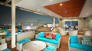 Um lounge no terraço com móveis de vime, teto de madeira e vista para a cidade à noite.