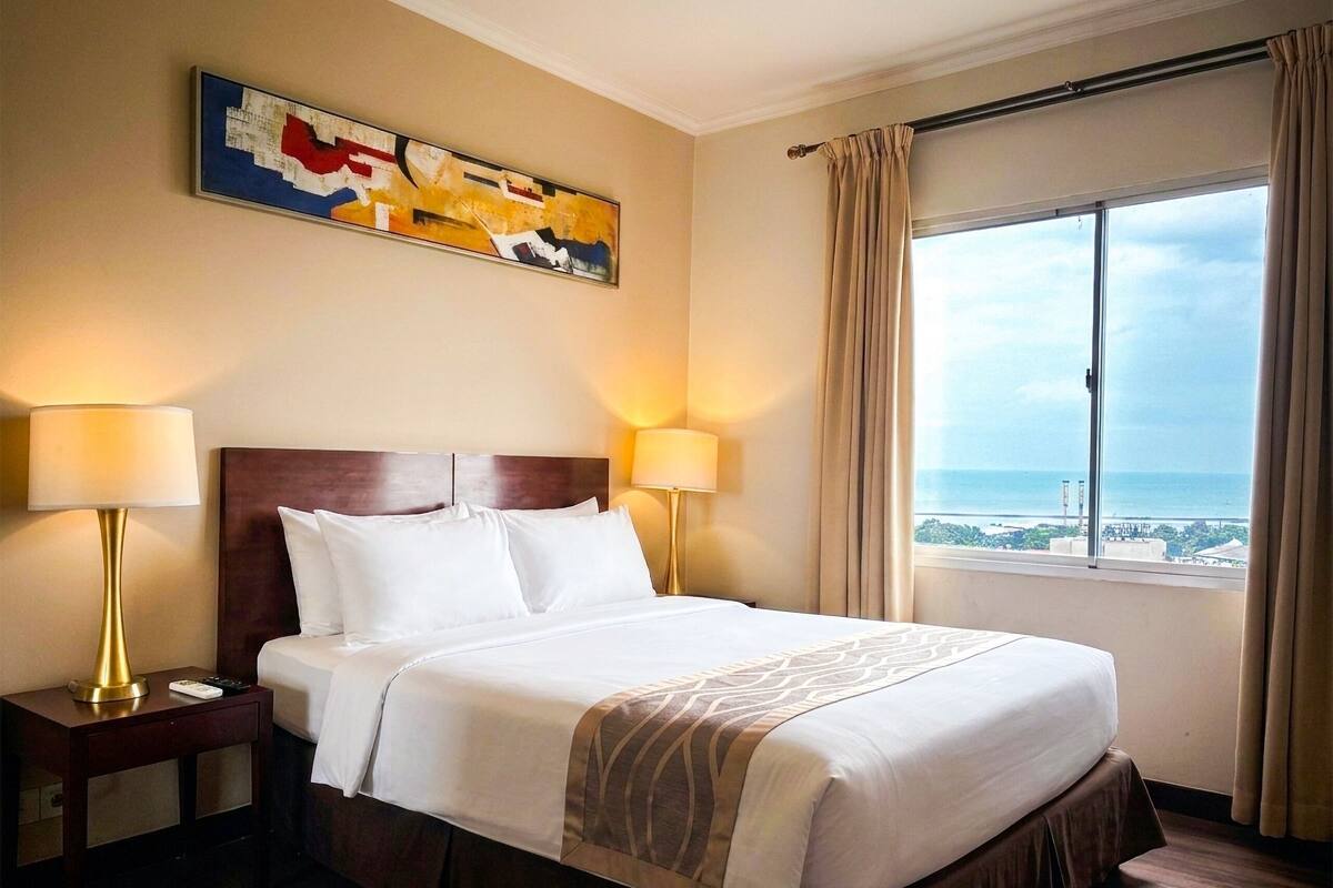 Une chambre d’hôtel avec un grand lit, deux lampes de chevet, un tableau au mur et une vue sur la mer par la fenêtre.