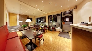 A modern hotel room with a wooden floor, a dining area with a table and chairs, a red sofa, and a kitchen area with a refrigerator and sink. - Kallmeyer (Berlin)