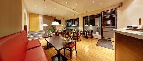 A modern hotel room with a wooden floor, a dining area with a table and chairs, a red sofa, and a kitchen area with a refrigerator and sink.