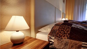A hotel room with a wooden nightstand, a lamp, a bed with patterned bedding, and a window with curtains. - Kallmeyer (Berlin)