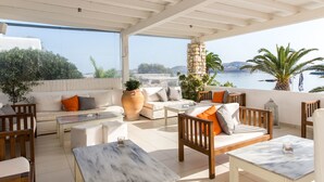 A sunlit outdoor seating area with a view of the sea, featuring a sofa, chairs, and a table. - Paros Bay Hotel (Paros)