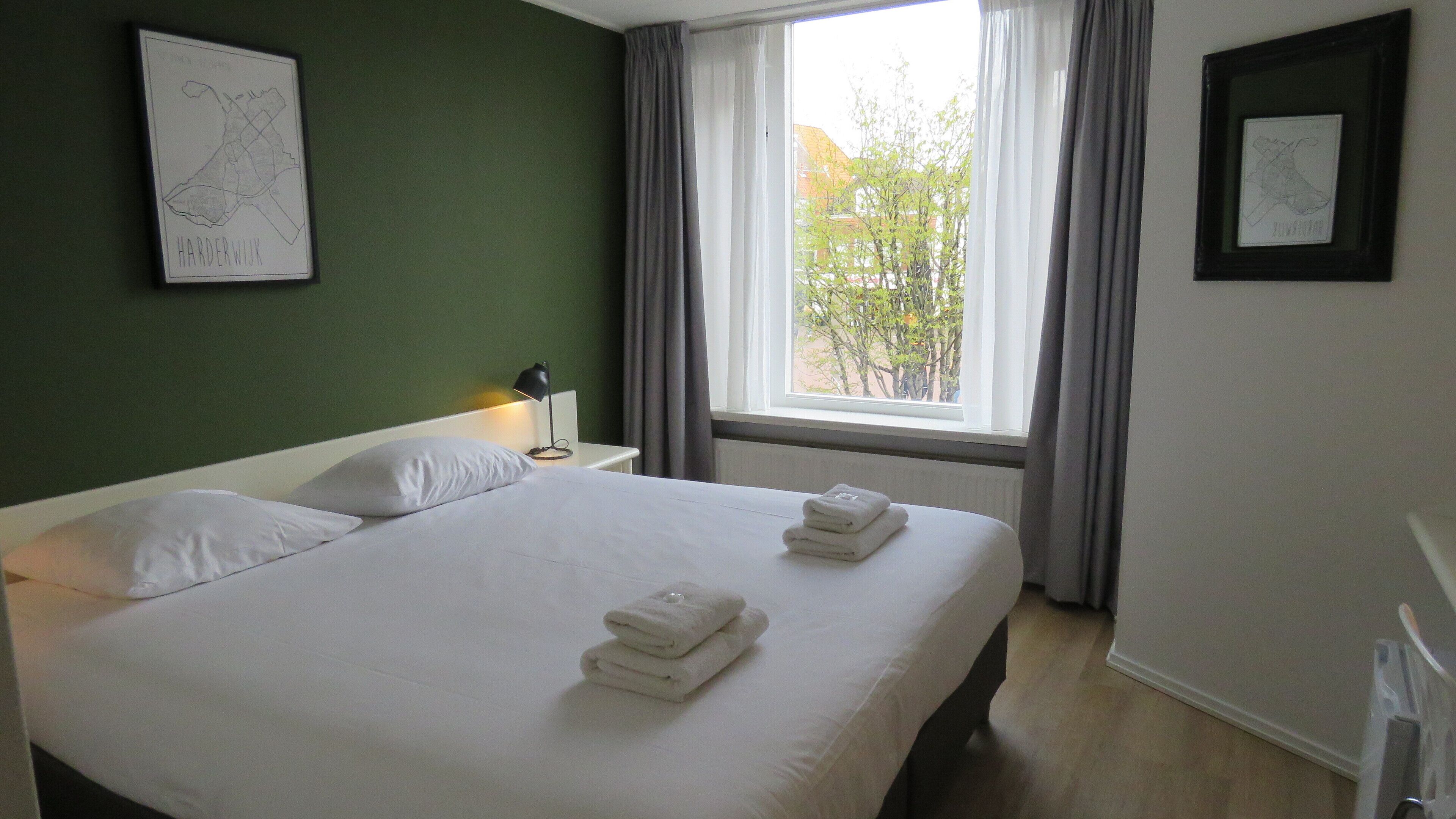 Camera d'albergo con un letto, un comodino, una lampada, una finestra con tende e una mappa incorniciata appesa al muro.