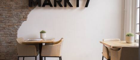 Un interior moderno con pared de ladrillo, pared blanca con señalización 'MARKT7', dos mesas de madera con sillas y plantas en macetas.
