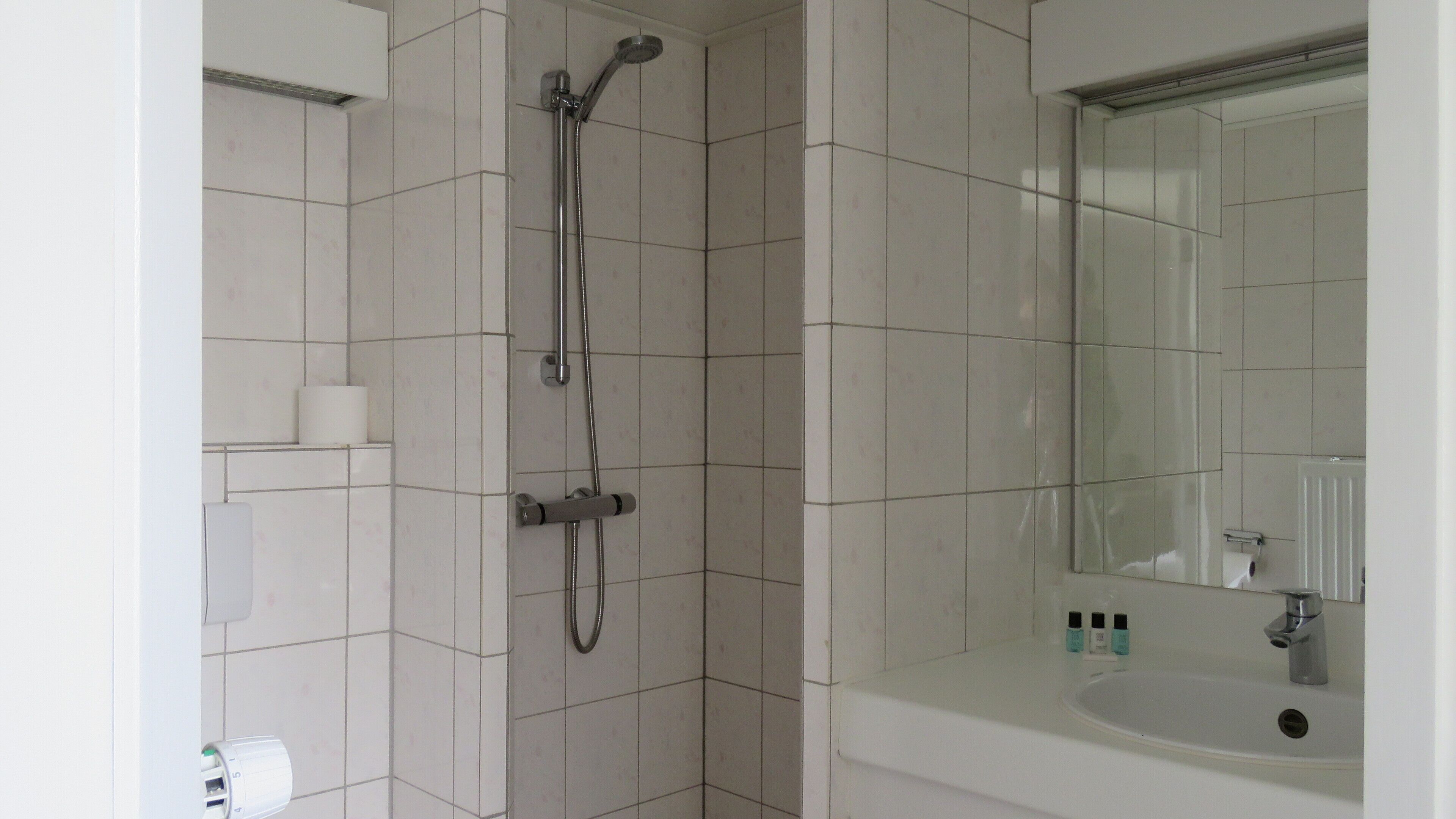 A bathroom with white tiles, a shower, a sink, and a mirror.