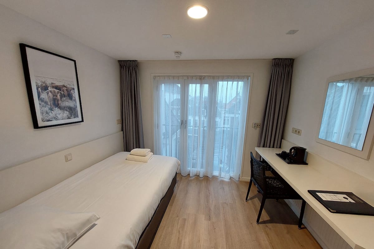 A hotel room with a bed, a desk, a chair, a window with curtains, and a framed picture on the wall.