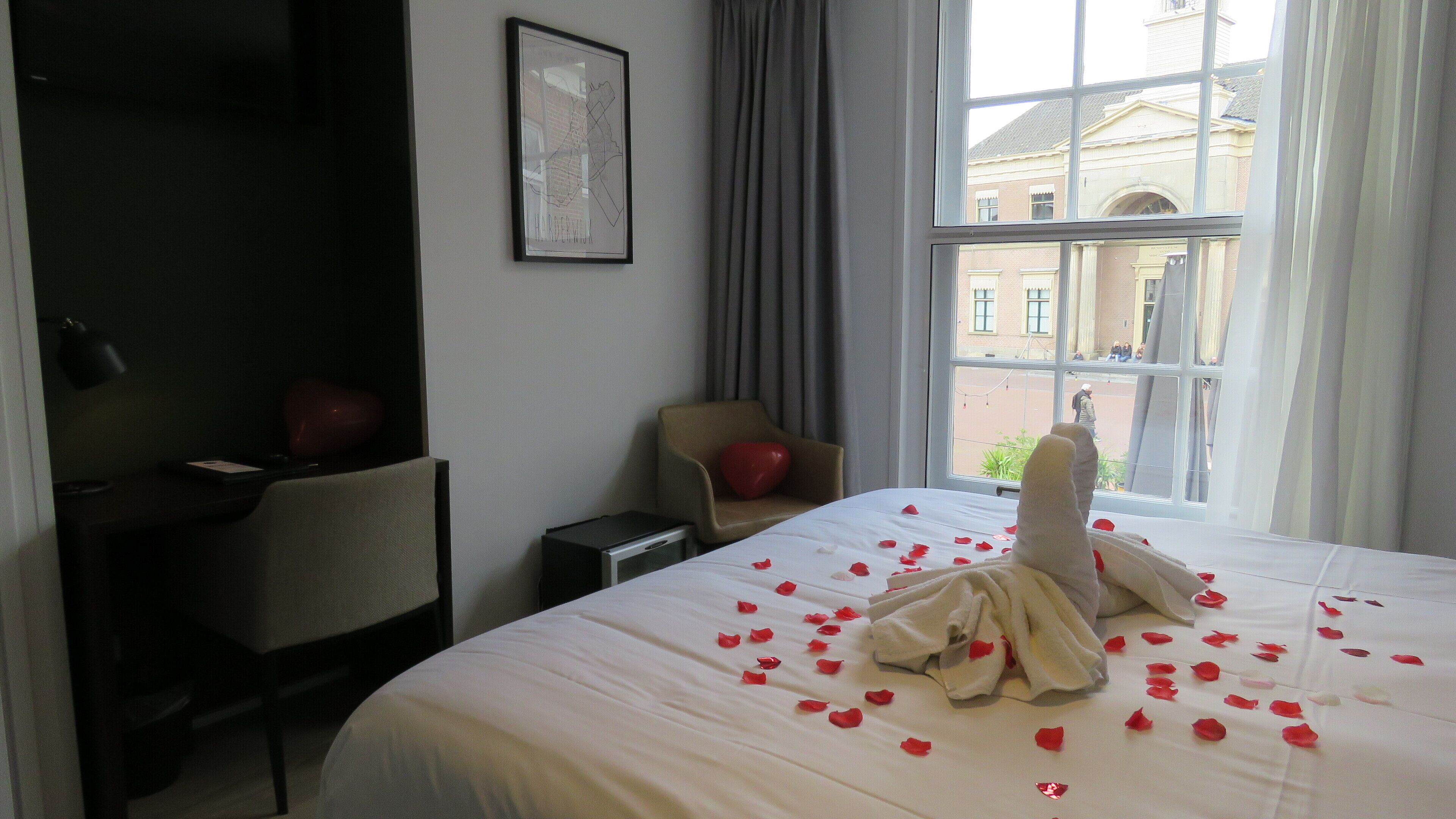 Habitación de hotel con una cama decorada con pétalos de rosa, un escritorio, una silla y vistas a un edificio a través de la ventana.