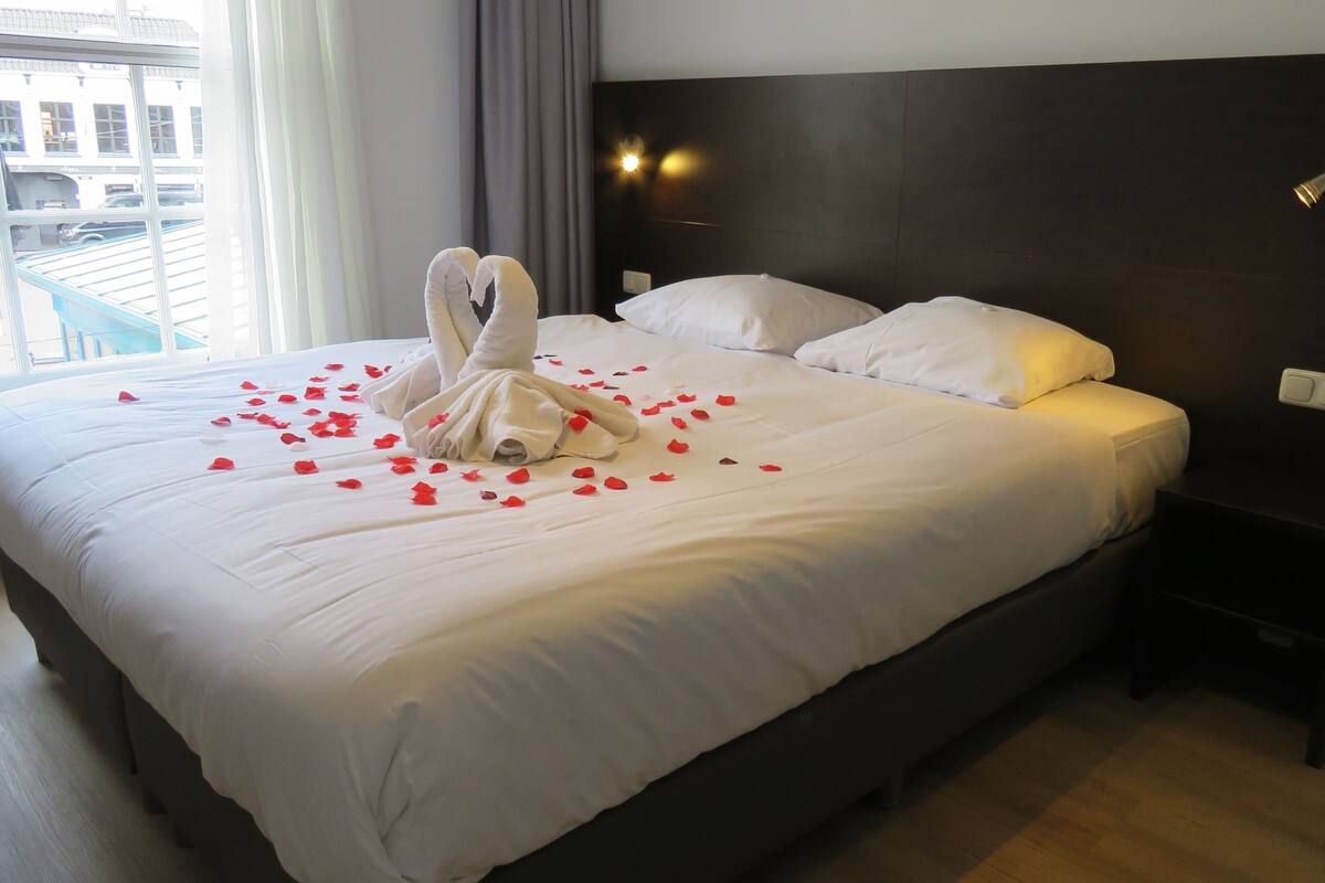 A hotel room with a neatly made bed, a headboard with wall-mounted lights, and a view of the outside through the window.