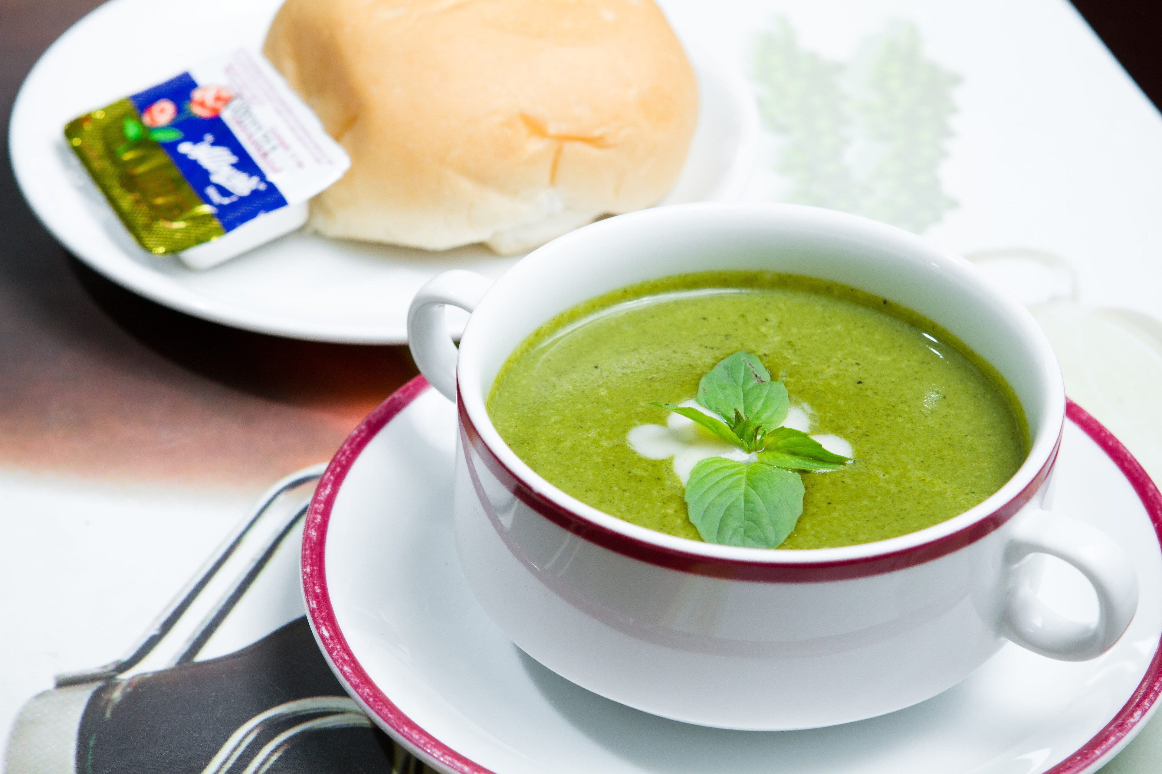Un tazón blanco con sopa verde y un borde rojo, un plato blanco con un panecillo y un paquete, y una cuchara sobre una mesa.