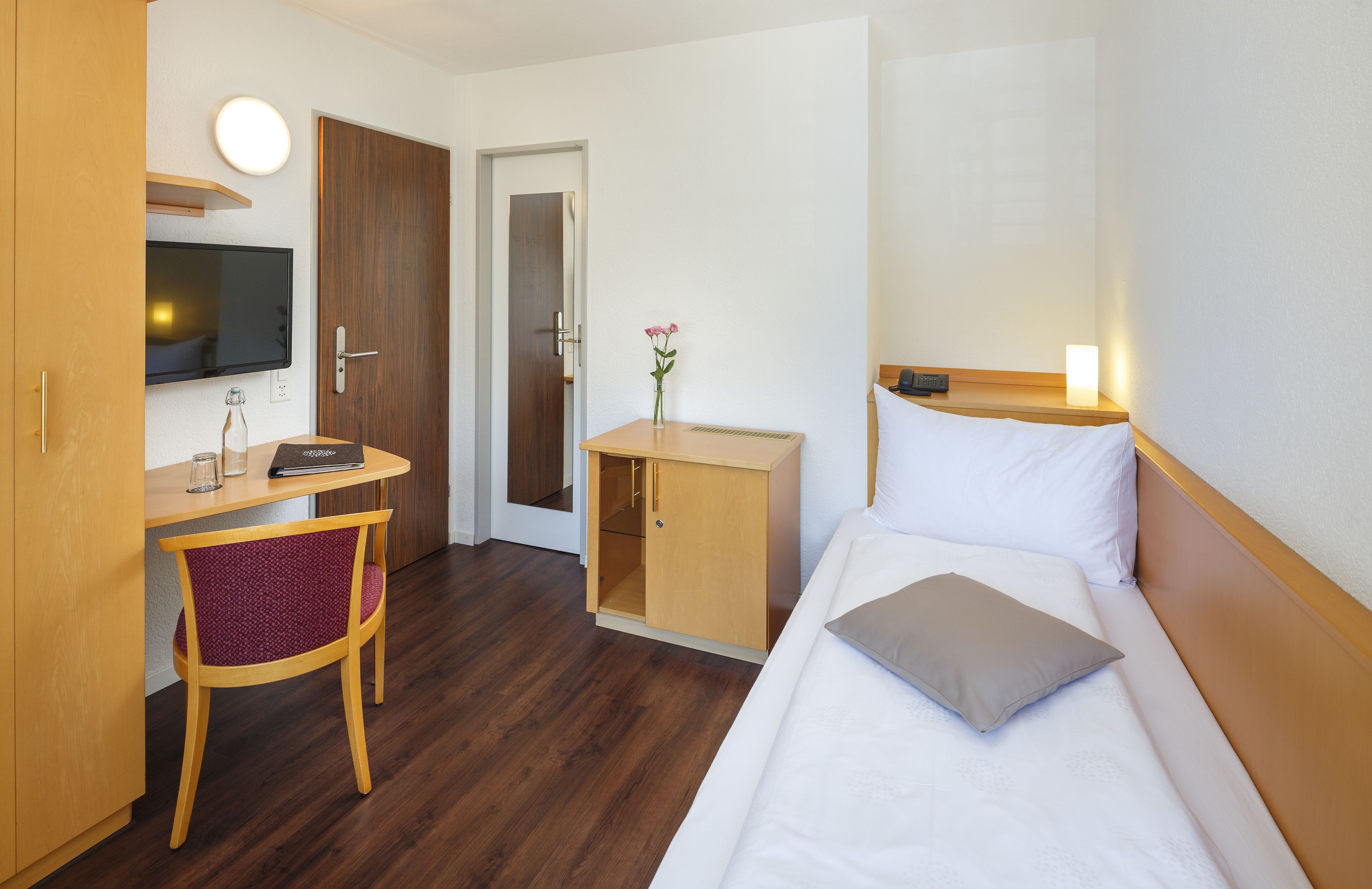 A hotel room with a bed, a desk with a chair, a television, a small cabinet, and a door.