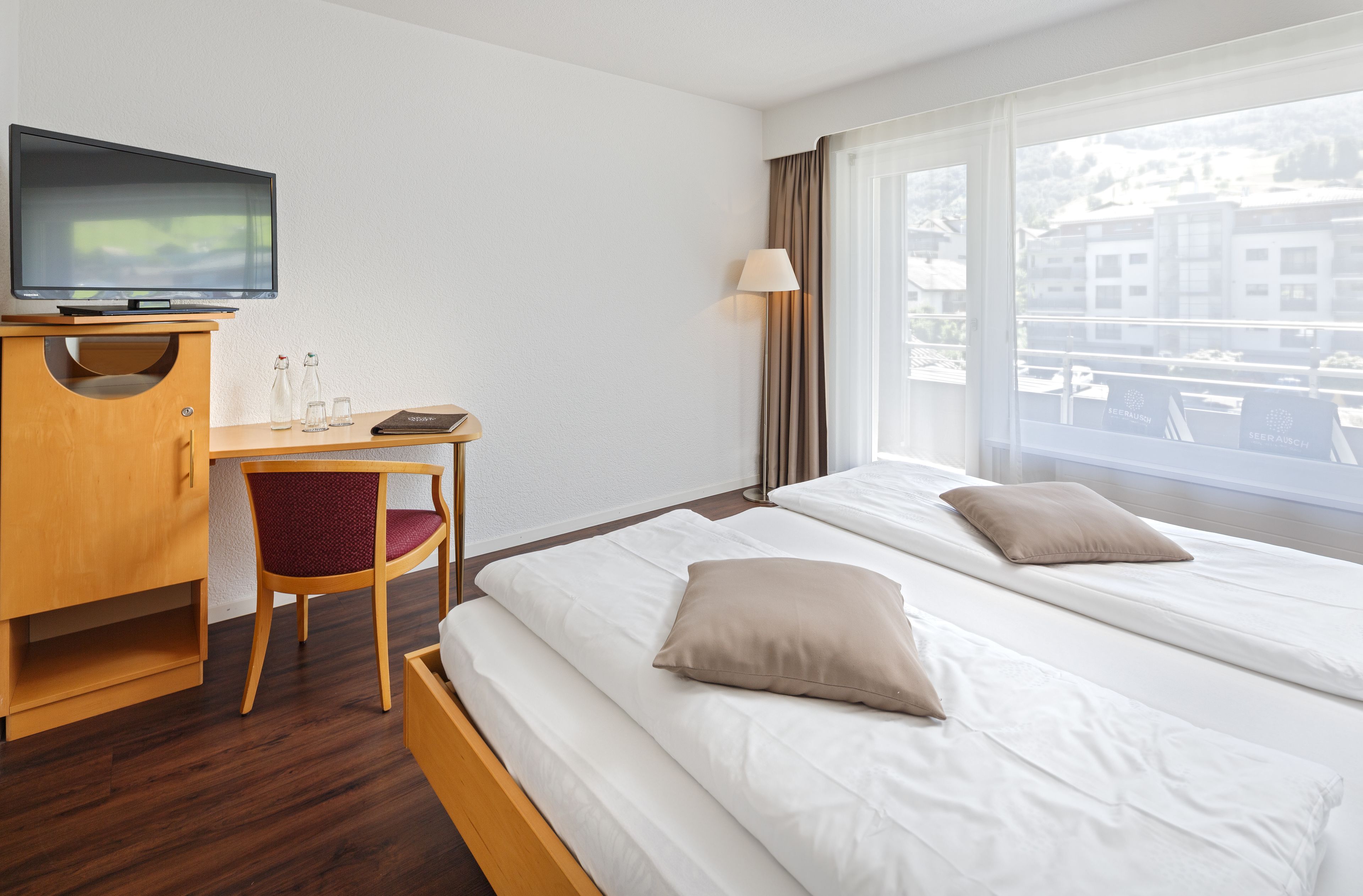 A hotel room with a bed, a desk with a chair, a television, and a window with a view of buildings.