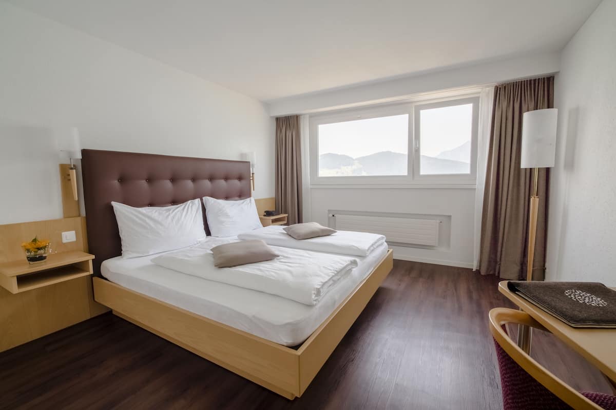 Een net opgemaakt bed met witte lakens en twee kussens, een houten nachtkastje met een lamp, een klein tafeltje met een bloevas, en een uitzicht op bergen door het raam.