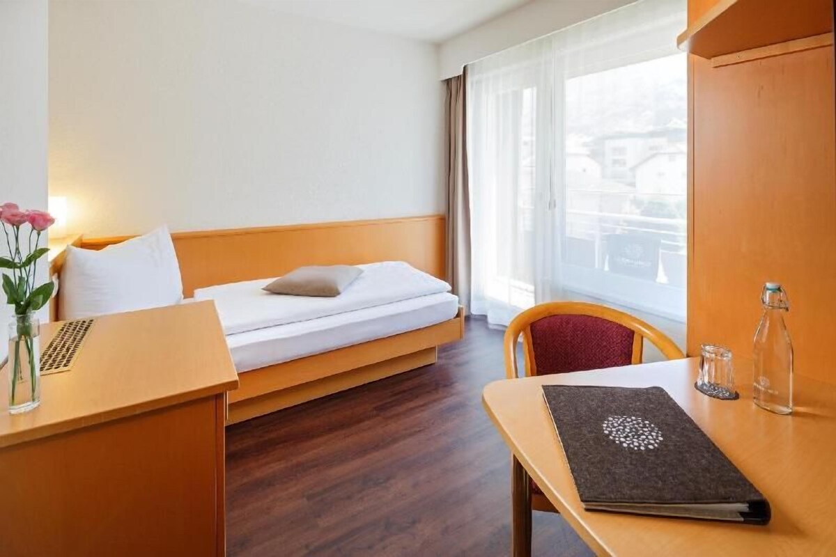 A hotel room with a single bed, a wooden desk, a chair, a small table, and a window with curtains.