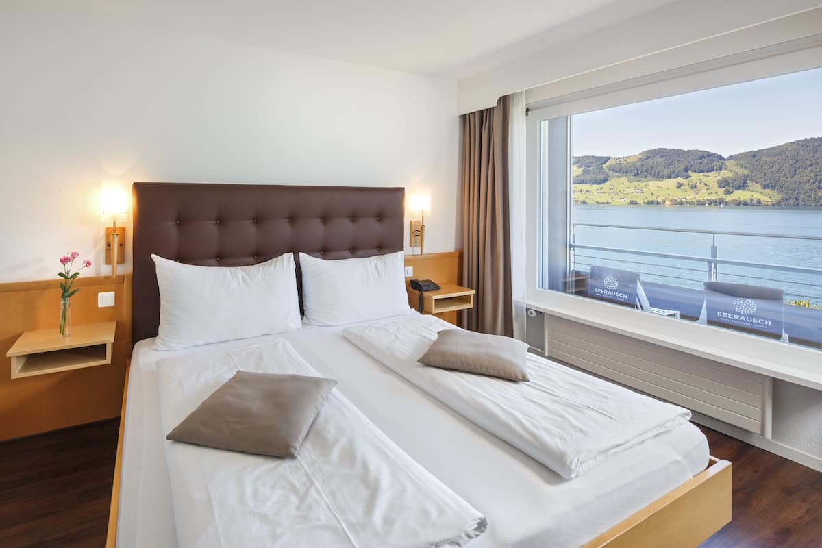 Een hotelkamer met een groot bed, een nachtkastje met een lamp, een raam met uitzicht op een meer en heuvels, en een houten vloer.