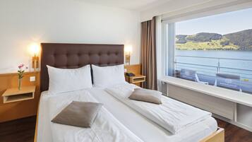 Ein Hotelzimmer mit einem großen Bett, einem Nachttisch mit Lampe, einem Fenster mit Blick auf einen See und Hügel sowie einem Holzboden.