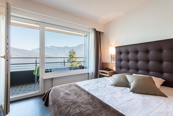 A hotel room with a large bed, a nightstand, a lamp, and a view of mountains through a sliding door.