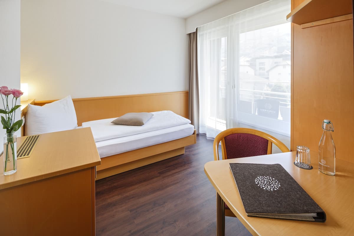Een hotelkamer met een bed, een bureau, een stoel, een vaas met bloemen, een fles en een raam met gordijnen.