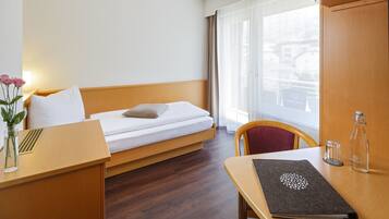 A hotel room with a bed, a desk, a chair, a vase with flowers, a bottle, and a window with curtains.