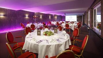 A banquet hall with round tables set for dining, red upholstered chairs, and a purple-lit backdrop.