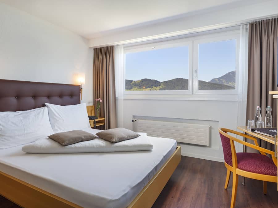 A hotel room with a bed, a desk, a chair, a television, and a window with a view of hills.