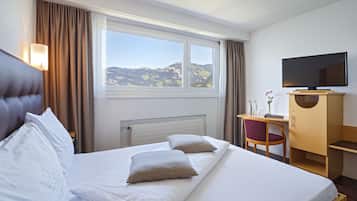 Une chambre d’hôtel équipée d’un lit, d’un bureau, d’une chaise, d’une télévision et d’une fenêtre donnant sur des collines.