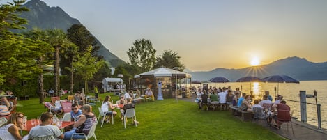 Zona de estar al aire libre frente al lago, con mesas y sillas, rodeada de vegetación y con montañas al fondo.
