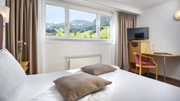 A hotel room with a bed, a desk, a chair, a TV, and a window with a view of green hills.