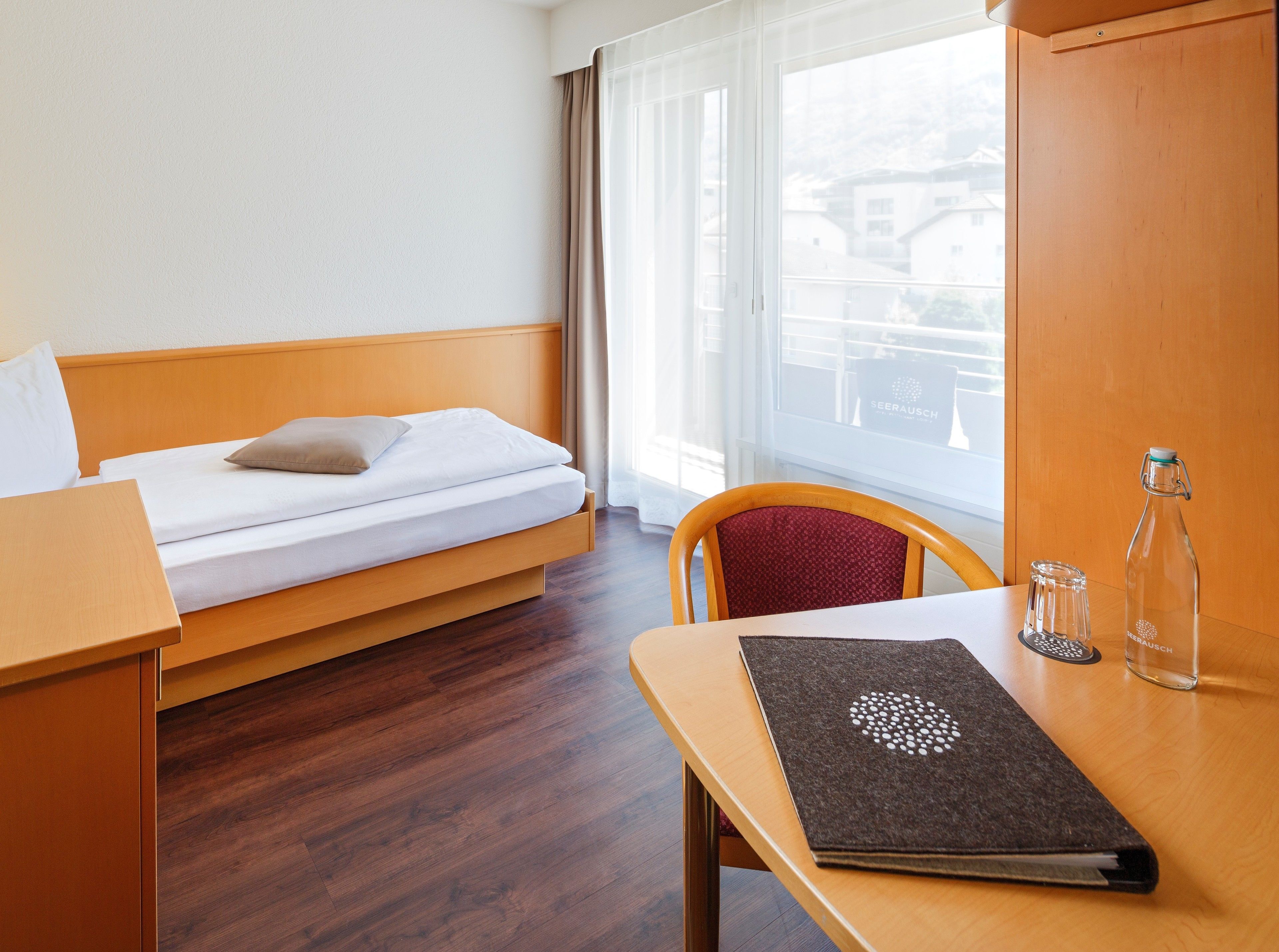 A hotel room with a bed, a wooden table, a chair, a window with curtains, and a view of buildings outside.