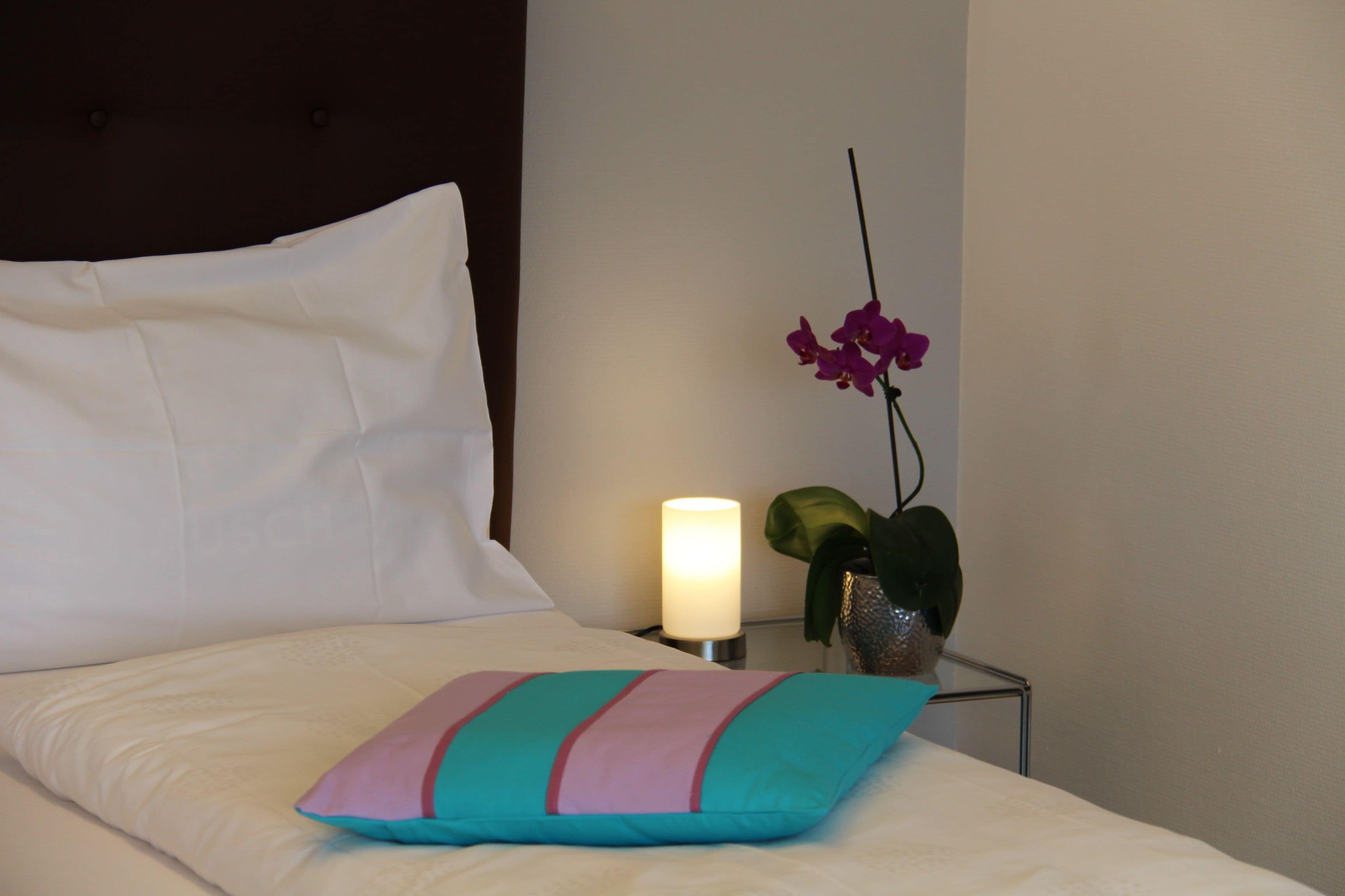 A neatly made bed with a white pillow and a colorful pillow, a bedside lamp, and a potted orchid plant.