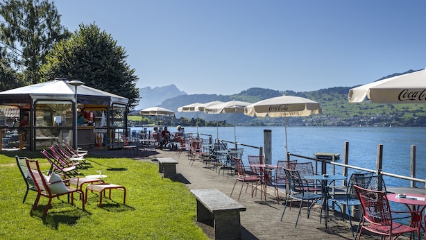 Ein Außenbereich mit Tischen, Stühlen und Sonnenschirmen, mit Blick auf den See.