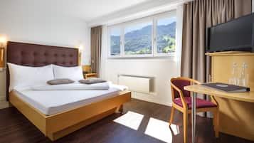 A hotel room with a bed, a desk, a chair, a television, and a window with a view of the mountains.