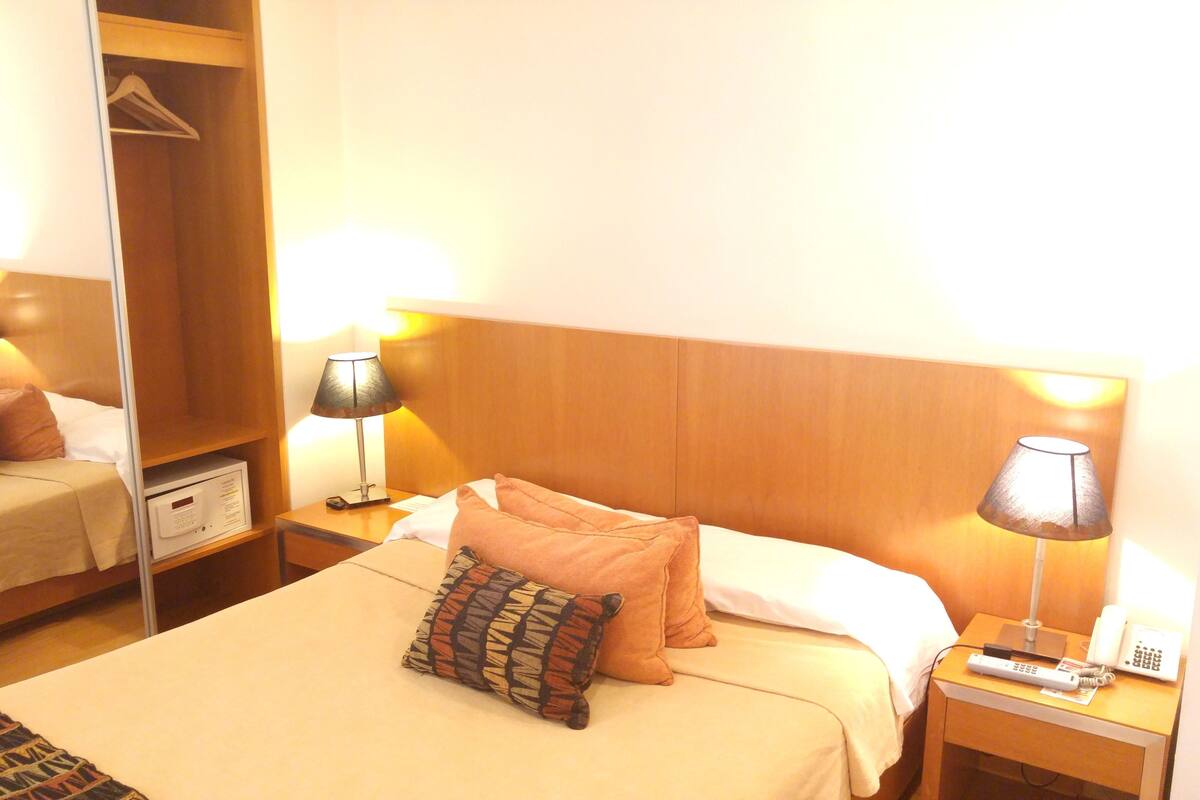 A hotel room with a bed, two bedside lamps, a bedside phone, a bedside table, a nightstand, a wardrobe, and a mirror.