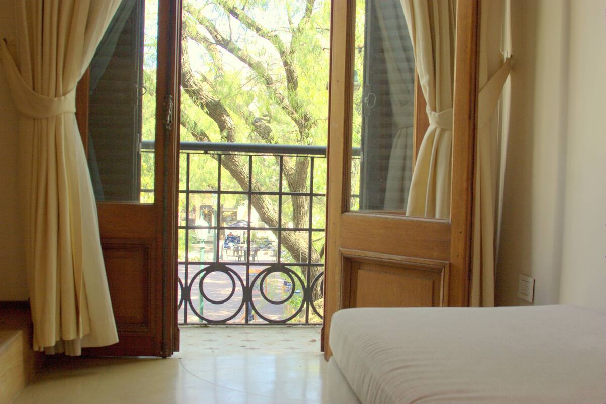 A room with a bed, a window with curtains, and a view of trees and a fence.