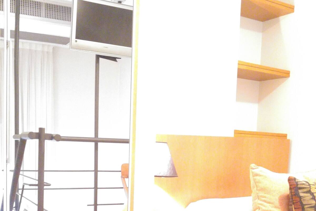 A hotel room with a bed, bedside table, and a desk with a chair. There is a window with blinds, a radiator, and a shelf above the bed.