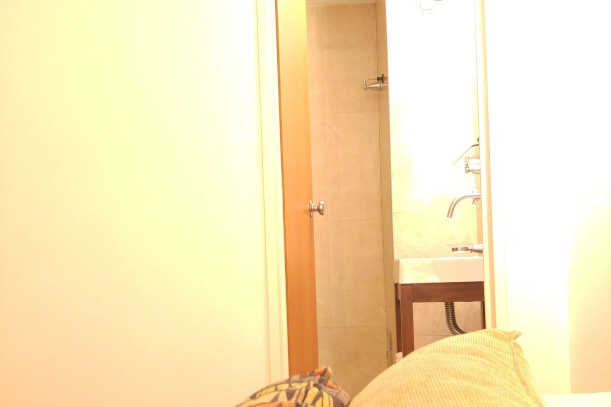 A hotel room with a bed, pillows, a door leading to a bathroom, and a mirror reflecting the bathroom.