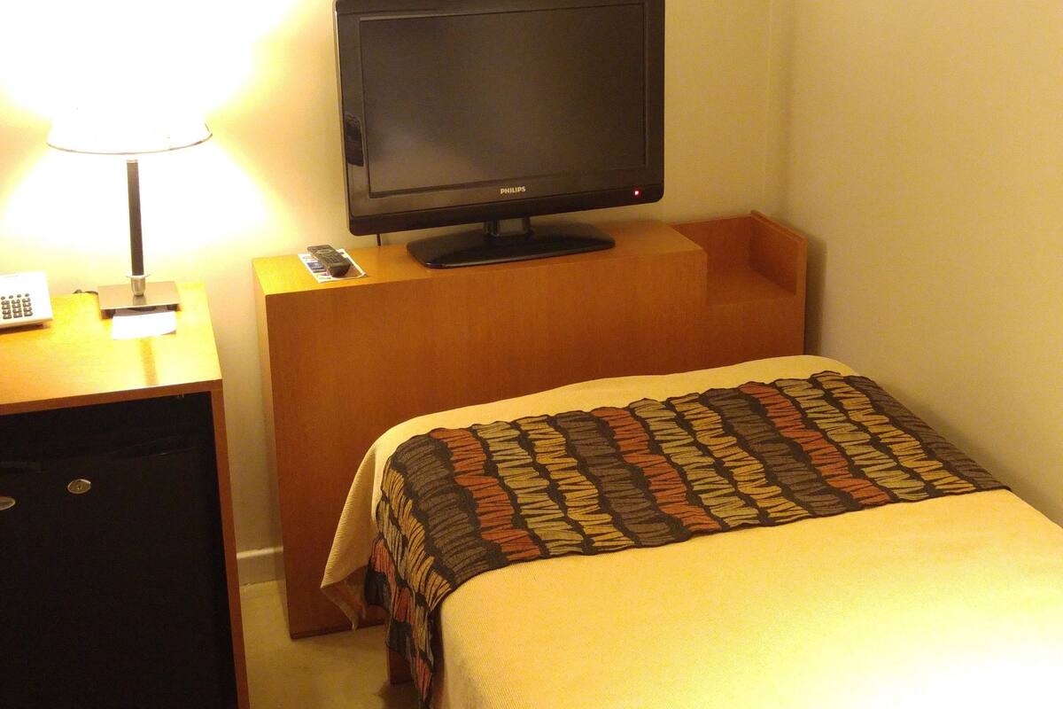 A hotel room with a bed, a television on a wooden stand, a nightstand with a lamp, and a small drawer.