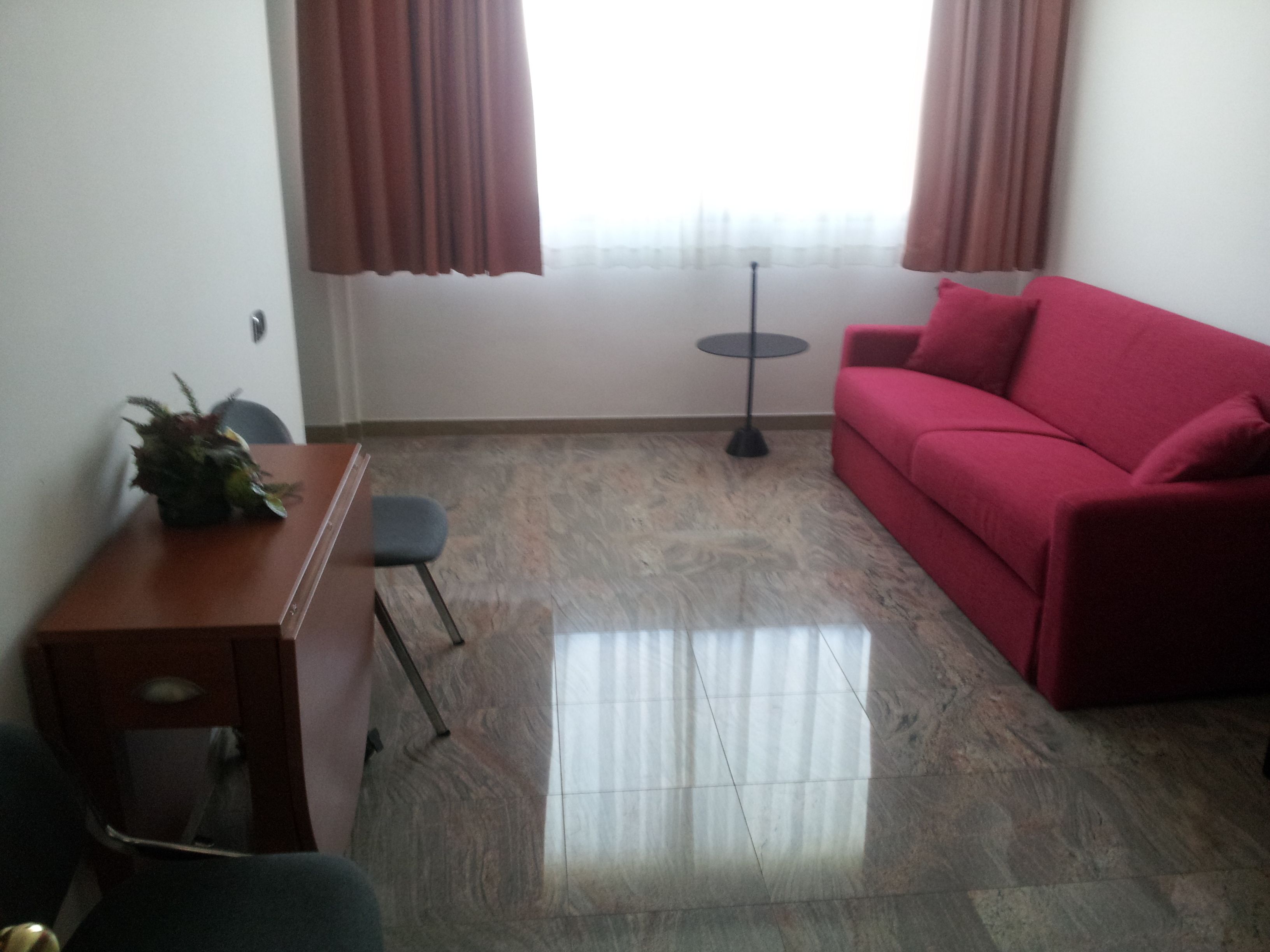 A room with a red sofa, a small desk with a plant, a chair, a round table, and a floor lamp.