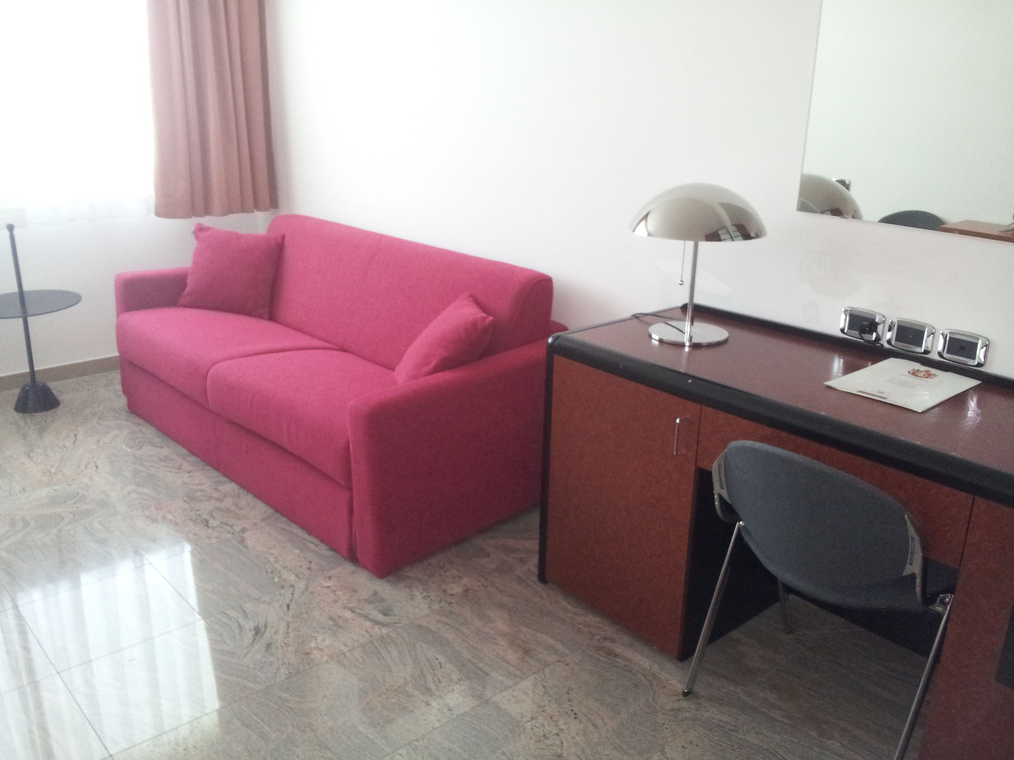 Una habitación con un sofá rojo, un escritorio con lámpara, una silla y un espejo.