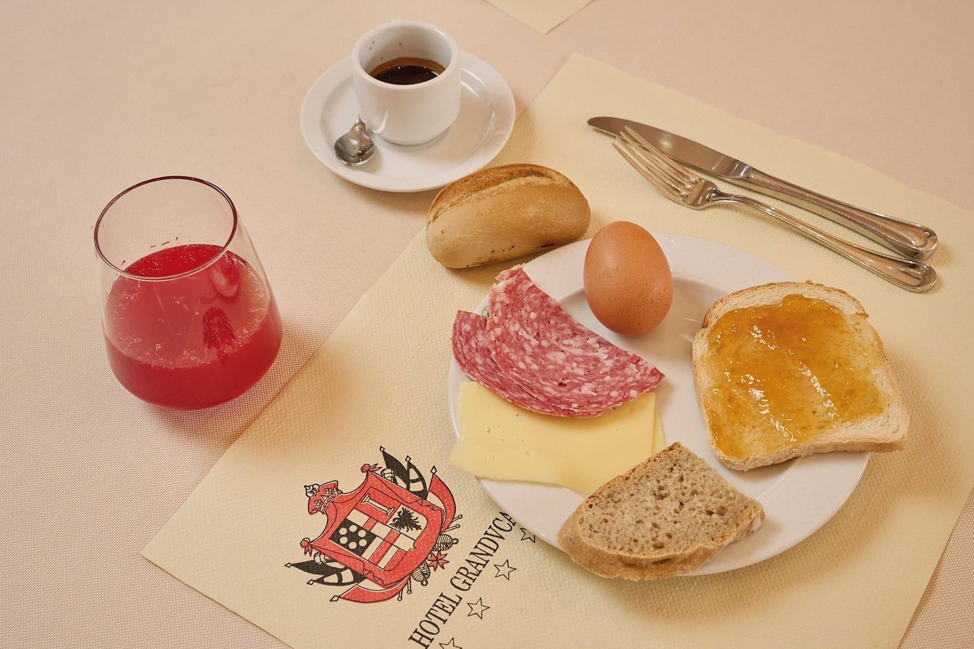 Piattino per la colazione con pane, formaggio, prosciutto, marmellata, un bicchiere di bevanda rossa e una tazza di caffè su un tavolo con tovagliolo e posate.