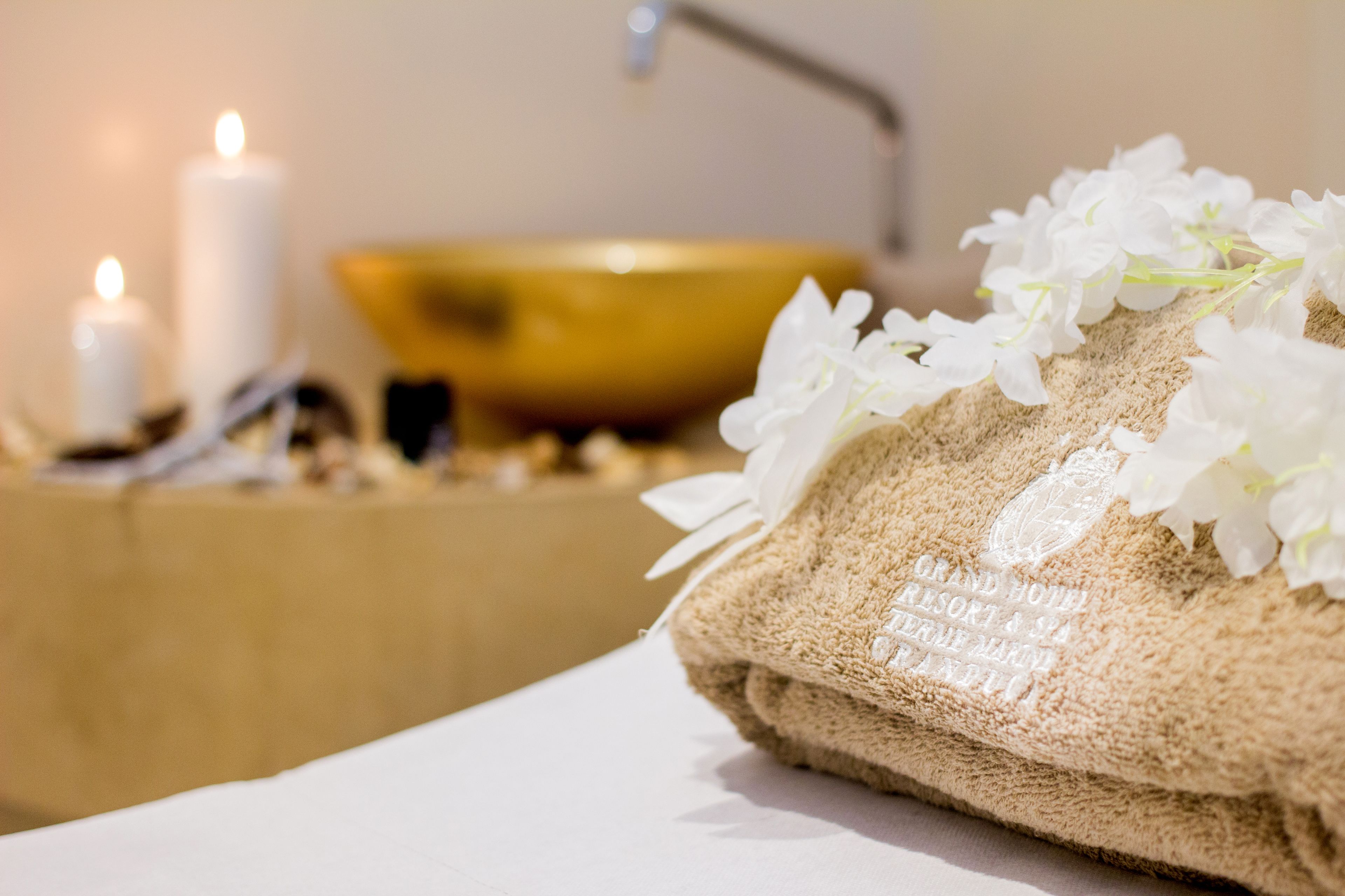 Ambiente de spa com toalha, flores brancas, velas e uma tigela dourada.