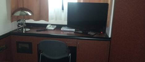 Habitación de hotel con escritorio, silla, televisor, teléfono y ventana con cortinas.
