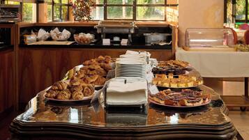 A buffet table with various pastries and desserts, a coffee machine, and a window with a view of greenery.