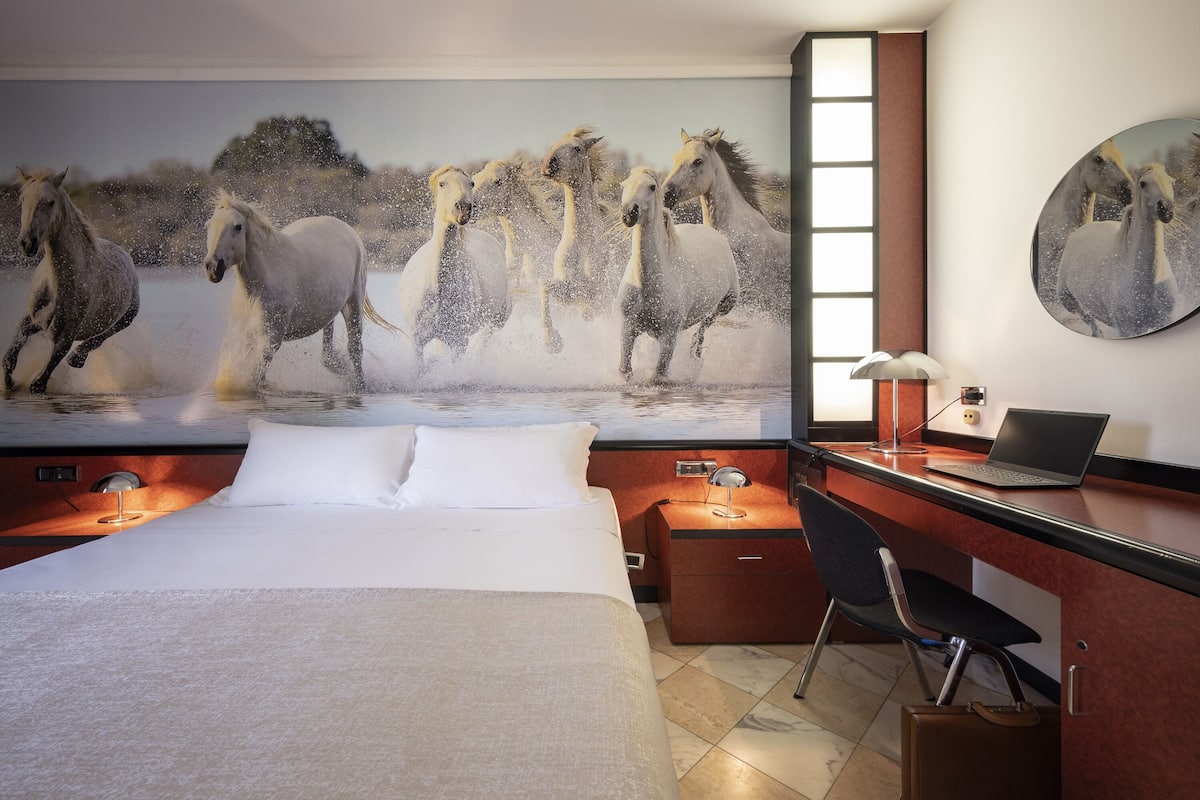 Hotelkamer met een groot muurschildering van paarden, een bed, een bureau met een laptop en een stoel.