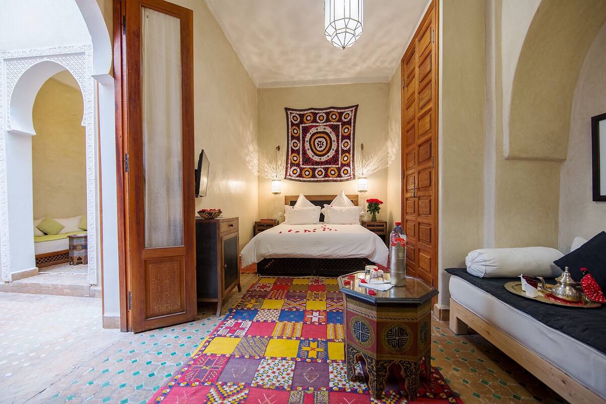 A bedroom with a colorful patchwork carpet, a bed with white linens, a wooden cabinet, and a wall tapestry.