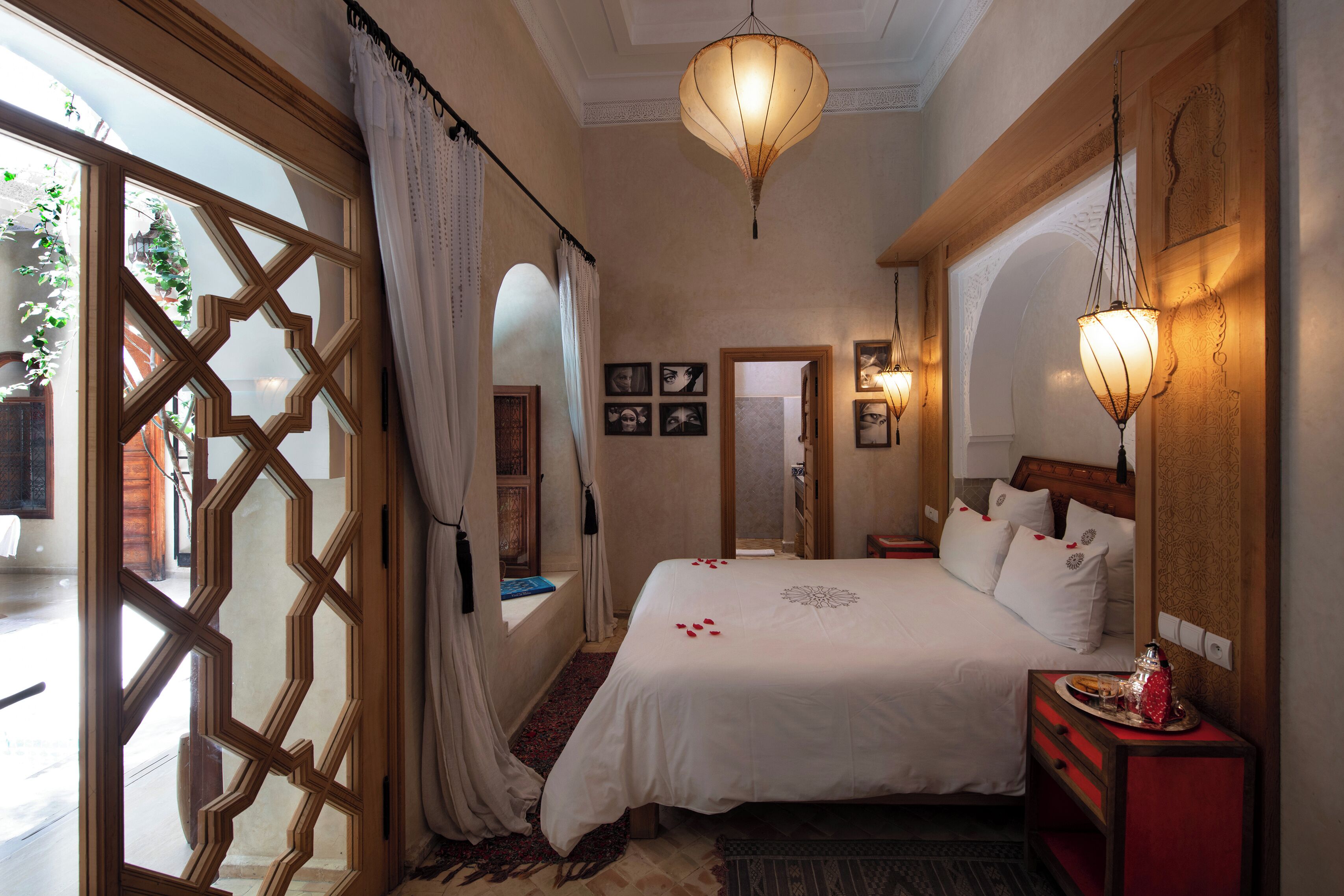 Une chambre d’hôtel dotée d’un grand lit, d’une tête de lit en bois, d’une table de chevet avec une lampe et offrant une vue sur l’extérieur grâce à une fenêtre dont la porte présente un motif géométrique.